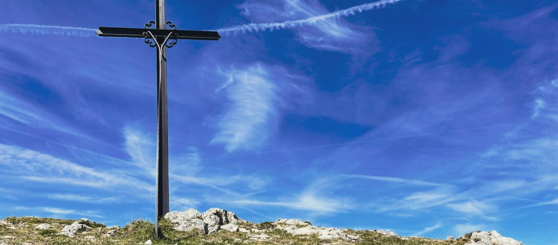 A photo of cross made of iron railings, set on a hill against a cloudy blue sky.