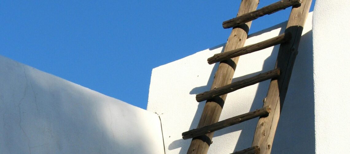 A photo of a native american lodging with white adobe walls, a wooden ladder, and a blue sky.